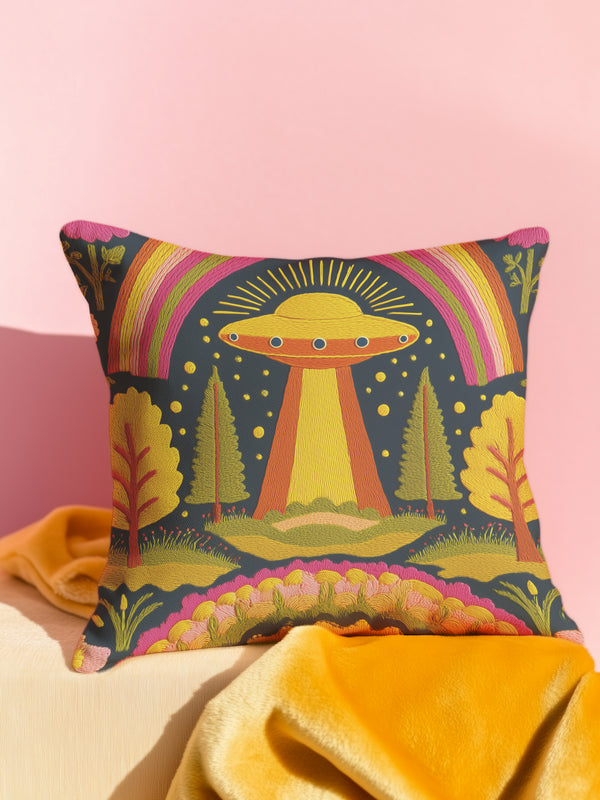 A colorful throw pillow with a whimsical alien design and various natural elements, such as trees and a rainbow, is displayed against a pink background.